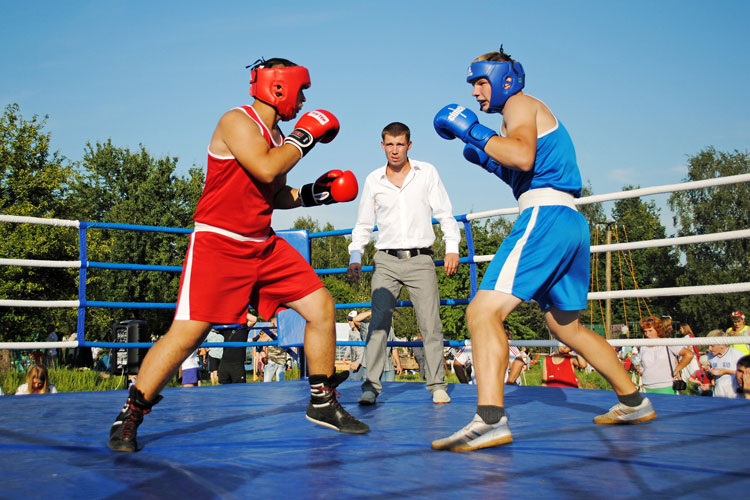 Boxhandschuhe kaufen: Du siehst zwei Boxer – einer in Rot, der andere in Blau – im vollen Kampfmodus, während der Schiedsrichter die Szene im Auge behält.