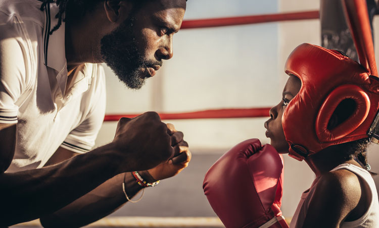 Engagierter Trainer im Gespräch mit jungem Boxanfänger mit roten Boxhandschuhen in einer Boxhalle, der die Grundlagen des Boxtrainings erlernt.