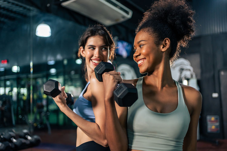 Zwei Frauen lächeln, während sie Hantelübungen im Fitnessstudio machen.