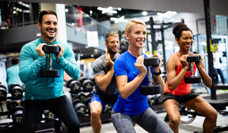Vier Personen beim Muskelaufbau-Training im Fitnessstudio, lächelnd mit Hanteln in der Hocke